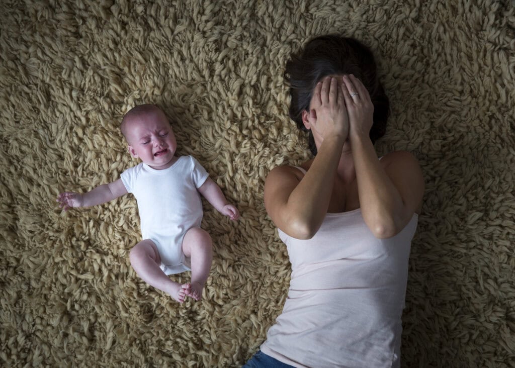 Mother lying on the floor crying next to crying baby, experiencing postpartum anxiety or postpartum depression and overwhelm. Therapy for perinatal mental health in NYC.