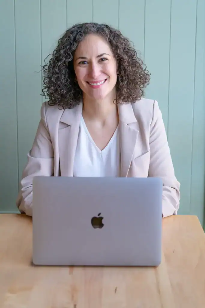 A smiling woman with curly hair sitting at a desk with a laptop, representing a professional therapist at Skye Ross Therapy, offering mental health support and counseling services.