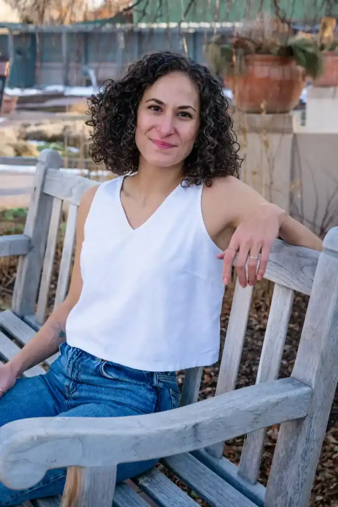 Portrait of Skye Ross, an EMDR therapist in NYC and Denver, sitting on a wooden bench outdoors, smiling, with a garden background, promoting mental health therapy for therapists, pregnant and postpartum people, and those living with chronic health conditions.